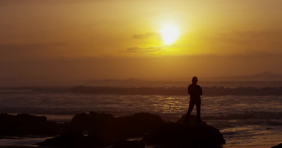 A person stands on a rock at the beach and looks out over the water