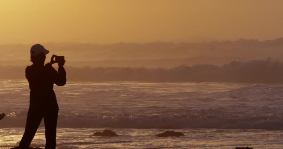 A person standing on a beach and sunset recording the tide