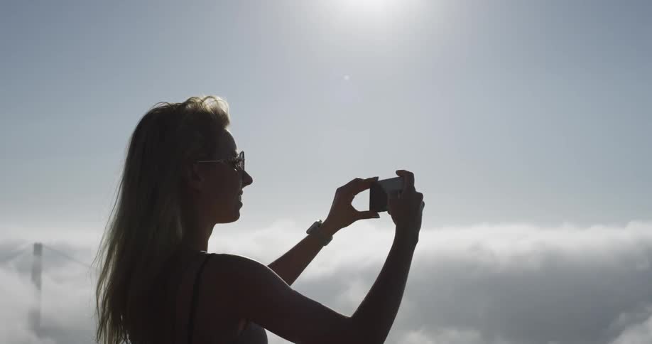 A woman takes photos on her cell phone of the fog and cloud covered bridge