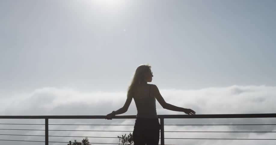 A silhouette of a woman walking up to a railing with clouds in the distance