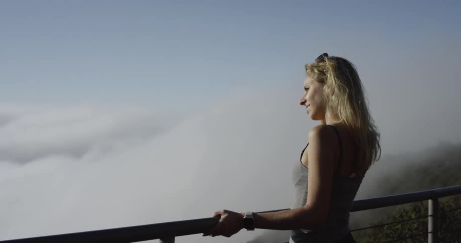 A woman stands at a railing to view the low clouds in a valley surrounded by hills