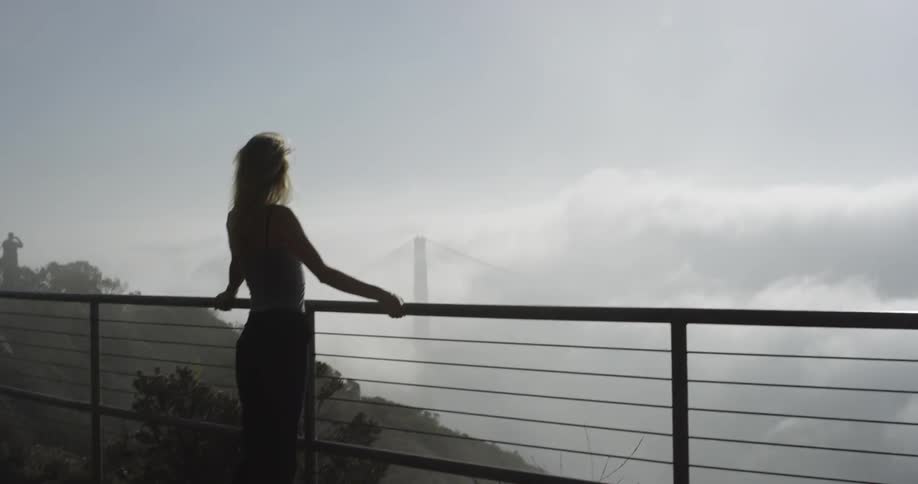 A woman stands at a fence on top of a hill that overlooks a fog enveloped suspension bridge