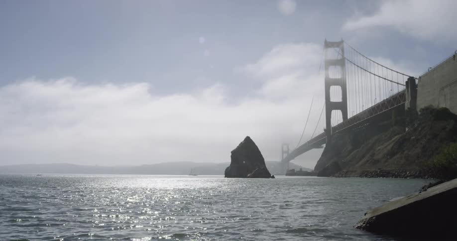 Large rock formation surrounded by rippling water near a cable bridge