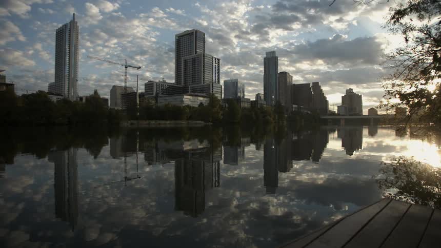 A city with a construction crane is reflected in a still body of water on a cloudy morning