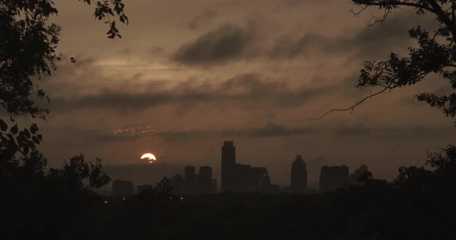 The sun is setting beyond a large city on a cloudy day