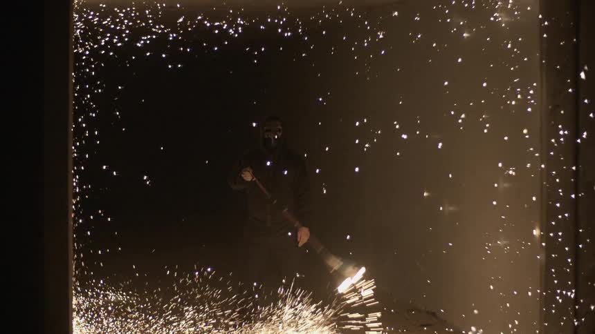 A person wearing a mask is swinging a flame in a circle as it shoots sparks everywhere