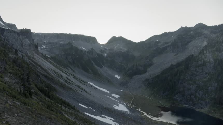 Rugged mountain plateau drops into a tree lined valley with alpine lake and melting snow fields