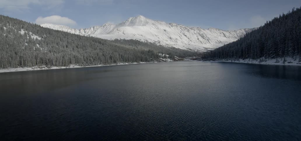 A serene river is lined by forest covered hills and leads to a snow covered range of mountains in the distance