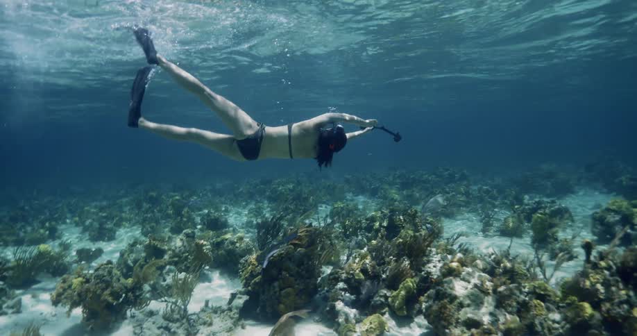 A woman swims in shallow open waters while wearing a snorkel and flippers and holding a selfie stick