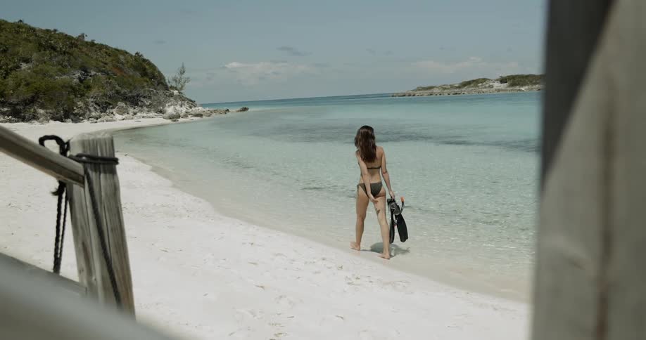 A woman walks on a sandy beach holding flippers and a snorkel