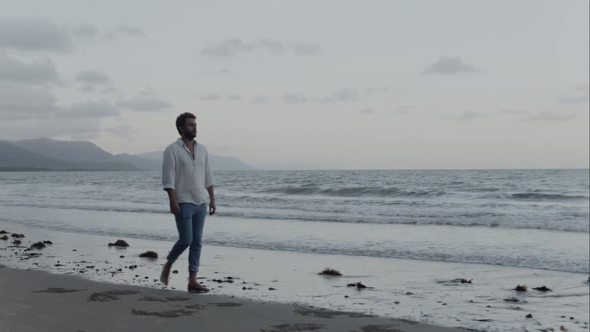 A man wearing jeans and shirt walks barefoot on the beach as waves come to shore at sunset