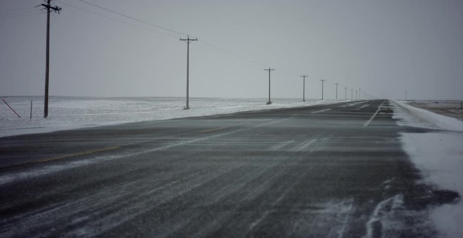 Wind blows snow across an empty rural road lined with telephone poles