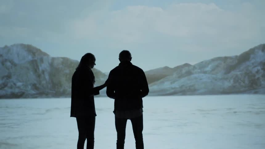 A man and a woman stand by a lake surrounded by snow covered mountains