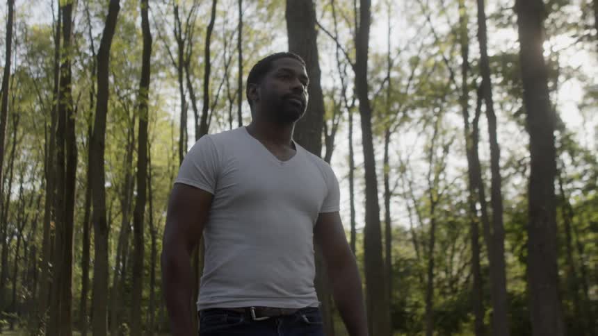 The sun peaks through leaves of trees in a forest as a man in a white shirt and jeans walks through