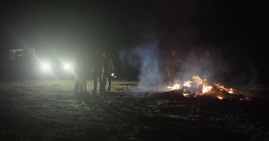 People are standing around a lit campfire at night