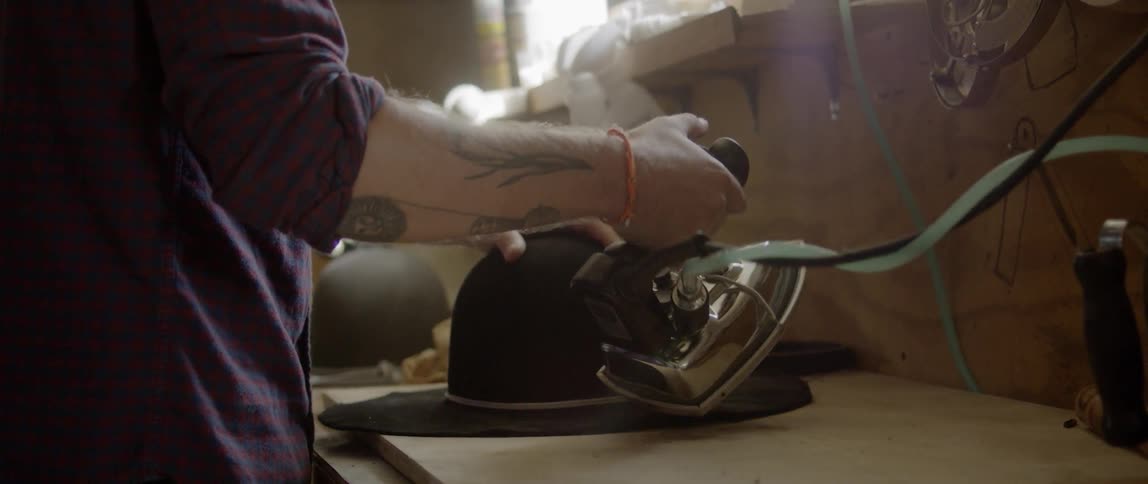 A white man uses an iron to flatten the brim of a hat
