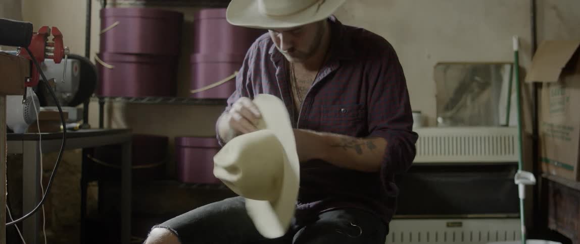A man sits and hand sews a hat