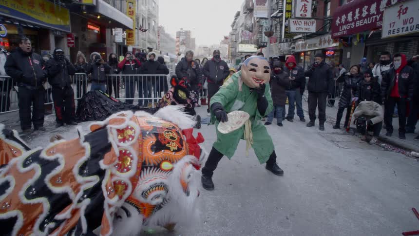 Performers in costume dance in a city street while an audience watches in the background