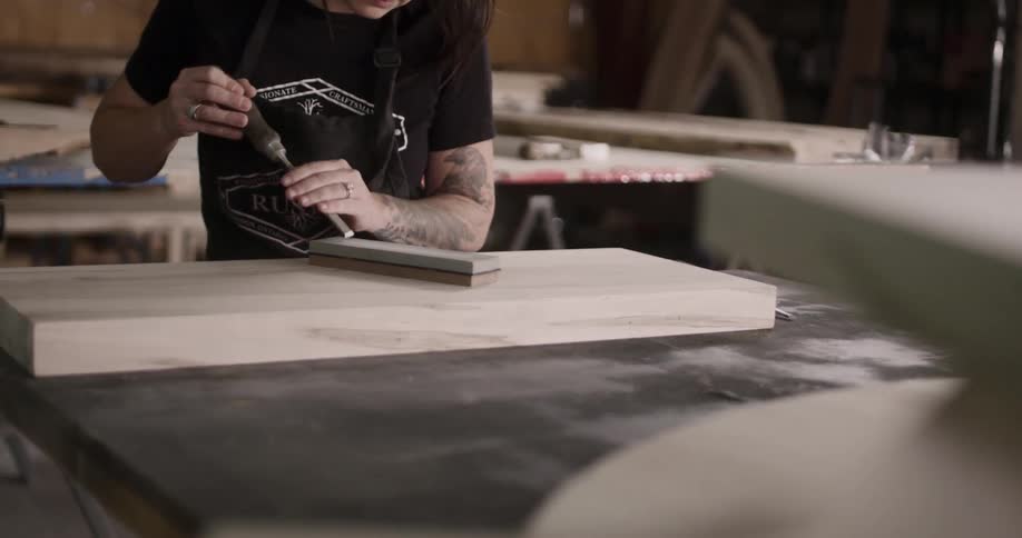 A white male with arm tattoos scrapes a piece of wood with a hand tool