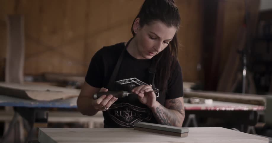 A white woman with arm tattoos scrapes a piece of wood with a hand tool