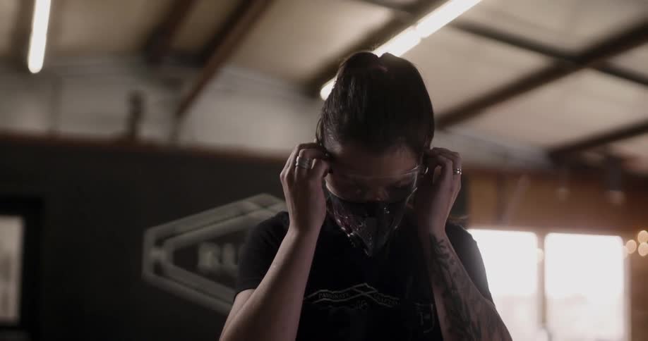 A woman with a tattooed arm puts on safety glasses in a workshop