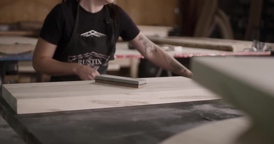 A woman works at a work bench with a chisel and piece of wood