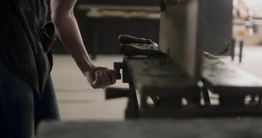 A person in a workshop wearing an apron turns the handle of a vice to more tightly hold a piece of wood