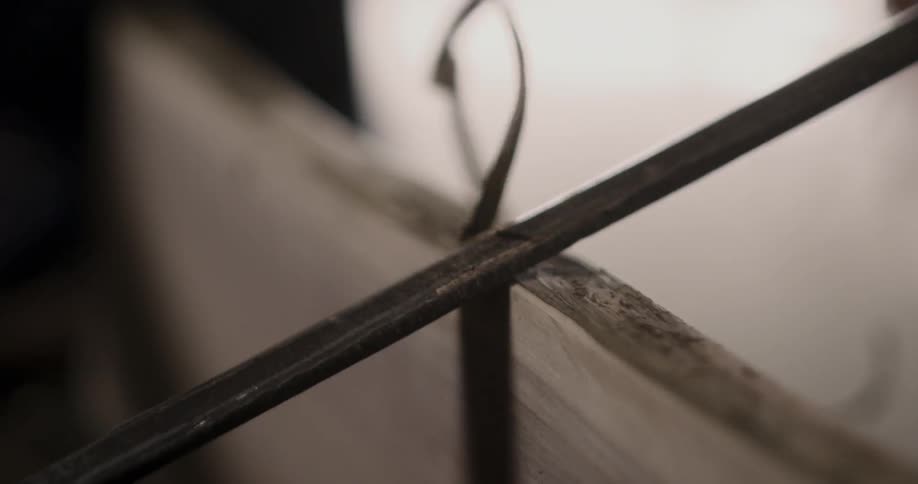 A white man scrapes a piece of wood with a metal hand tool