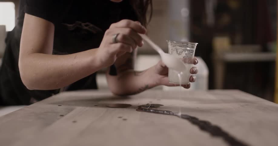 A woman using a spoon to drizzle oil over a dark liquid on a wooden surface