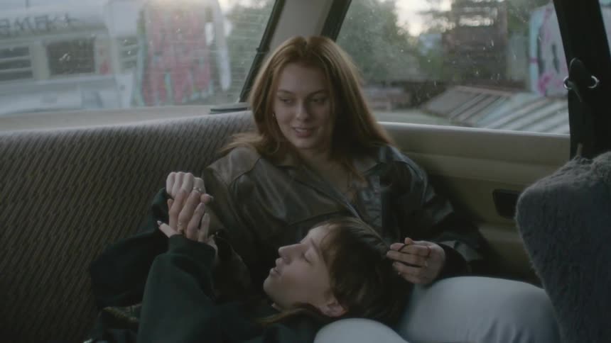 A young female is sitting the back seat of a car while her friend lays down in the back seat with her head on the leg of the other female