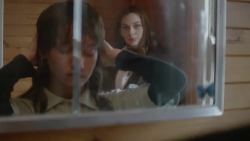 Woman adjusts her hair as she looks in a mirror as another woman watches