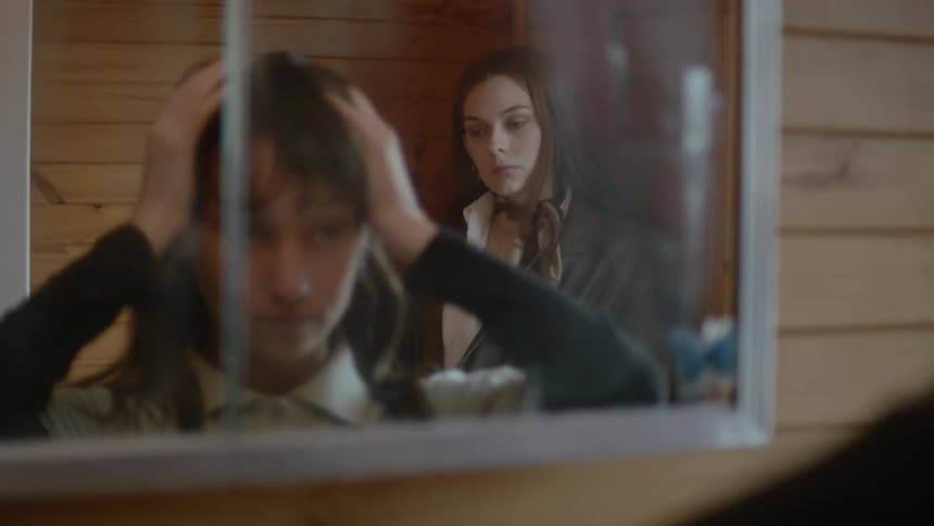 A young white girl fusses with her hair while looking the mirror while another white woman watches her in the background