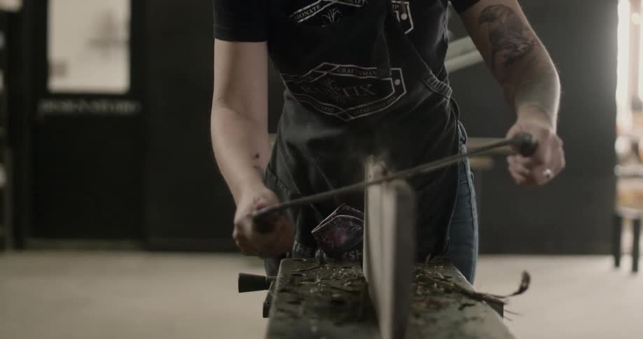 A man is using a dual handled tool to carve away at a piece of wood that is mounted on a work table