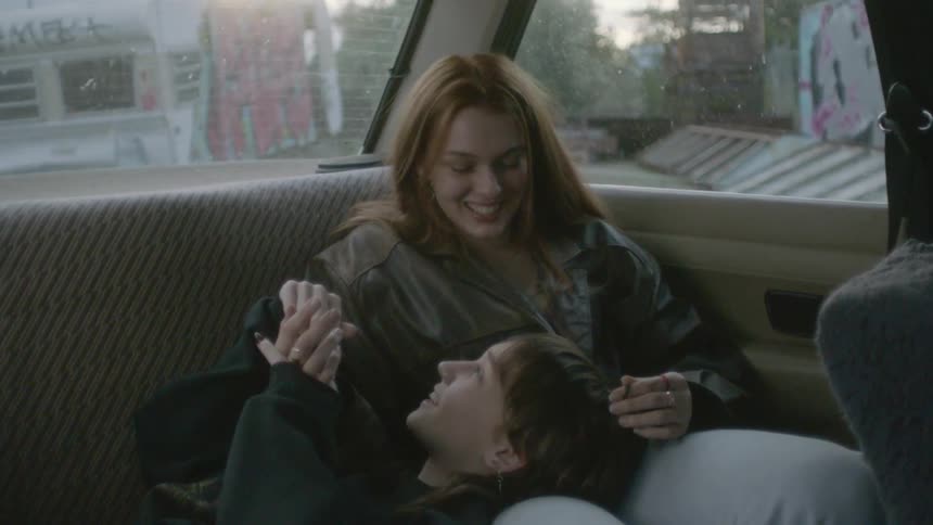 A teenage boy and a teenage girl sit in the back of a car with the boy resting his head on the girls lap