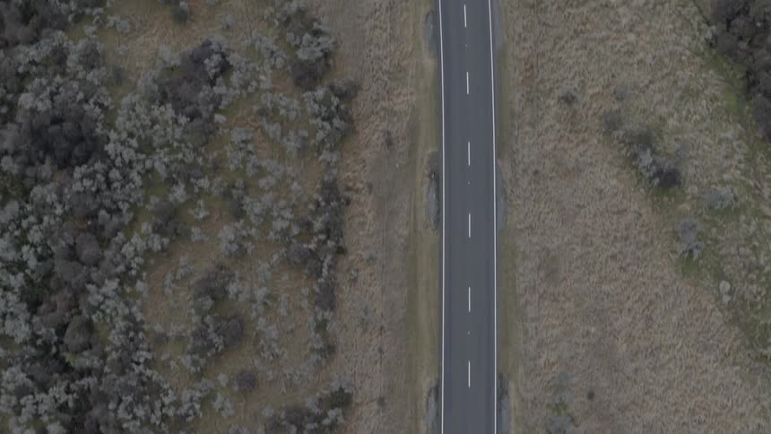 A car drives quickly along a two lane road that is carved through a sparsely forested landscape
