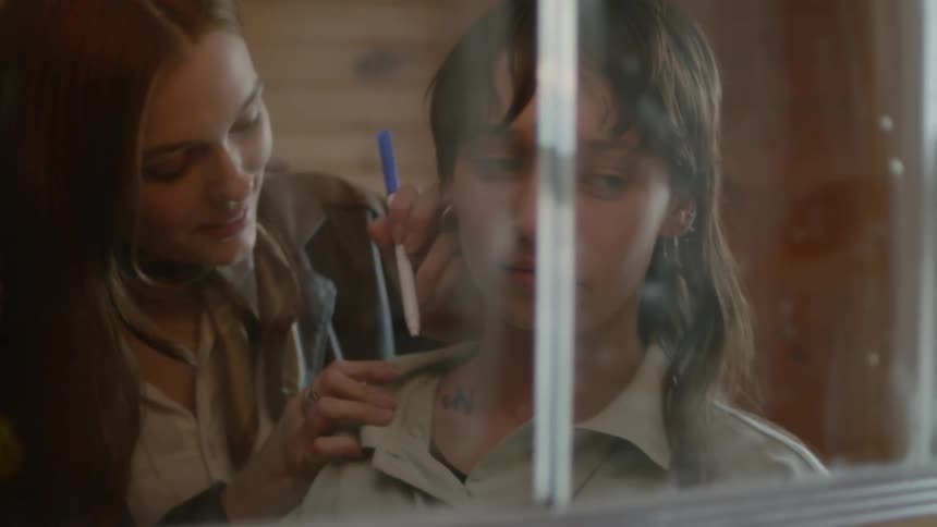 A woman uses a felt tip pen to write on the neck of her friend as they stand in front of a mirror in a wooden cabin