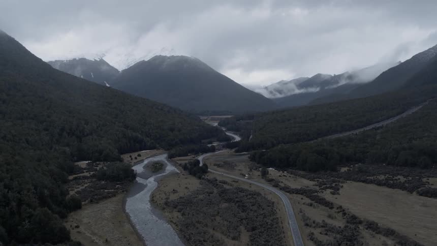 A road winds and curves up a forest covered hill and a stream flows down from the mountains