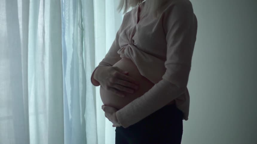 A pregnant woman rubs her stomach standing by a window