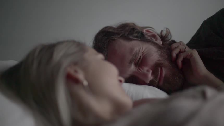 A blonde woman lying on a bed rubbing the beard of a man lying next to her