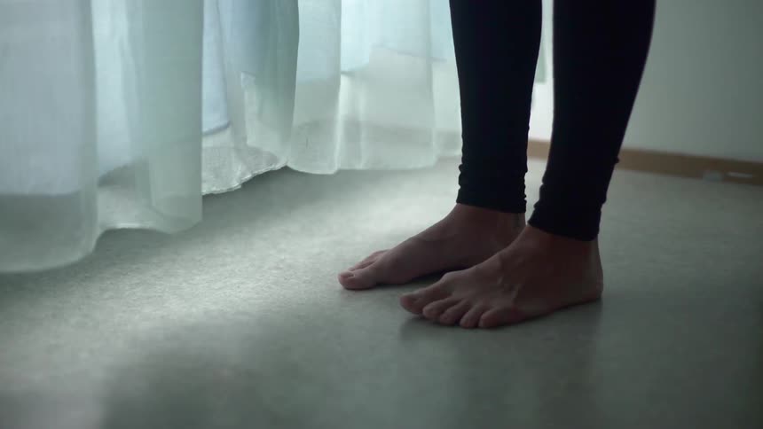 A woman stands barefoot in a room near a window with white curtains