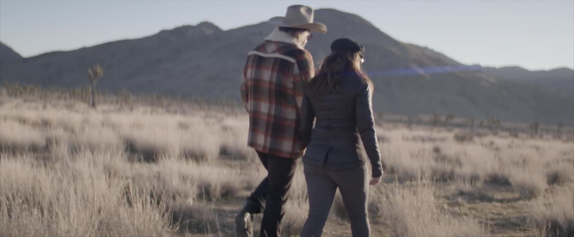 Woman and man wearing wide hat walk on dry grass by holding hands during daylight