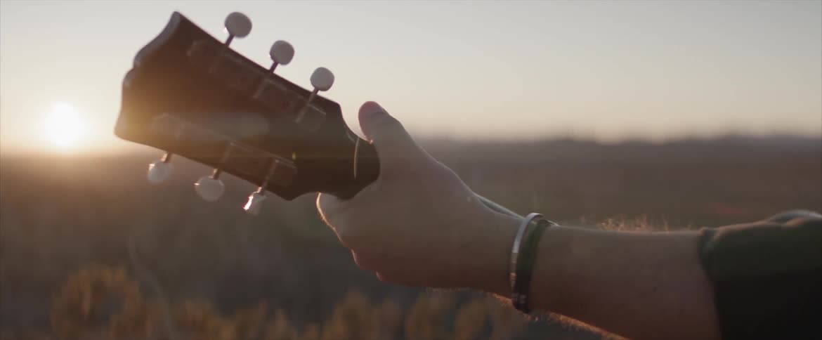 A man with bracelets on his wrist stands with an acoustic guitar outdoors at sunset