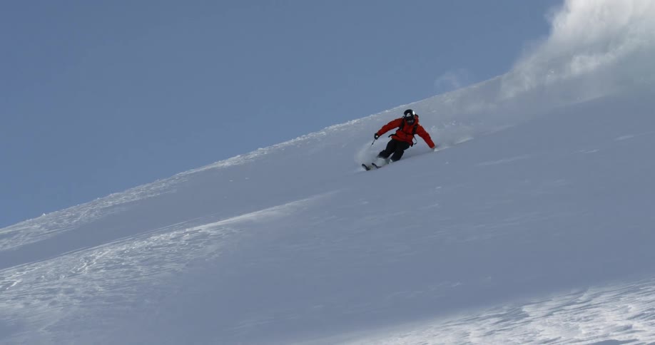 A skier in a red skijacket spins 360 degrees while skiing down a steep slope