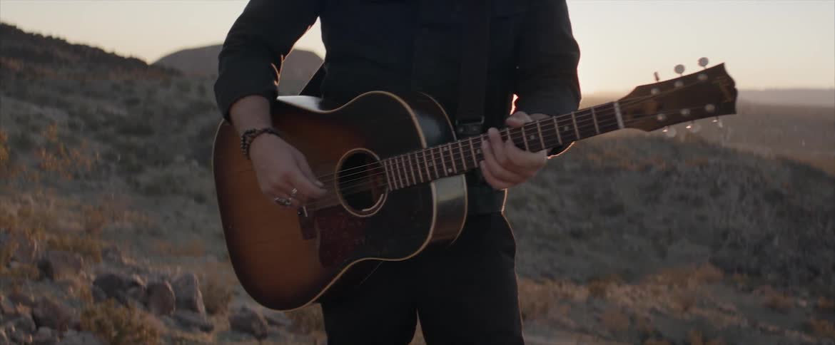 A man plays the guitar while standing outside in hill country
