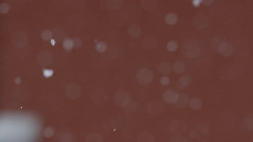 Small and lightweight snow flakes fall slowly downward to the snow covered ground in front of a red wall of a building