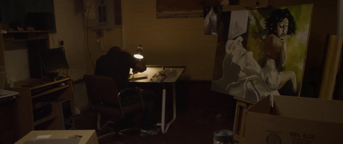 A person is sitting in a room with one light shining on him and art at a desk fixing something