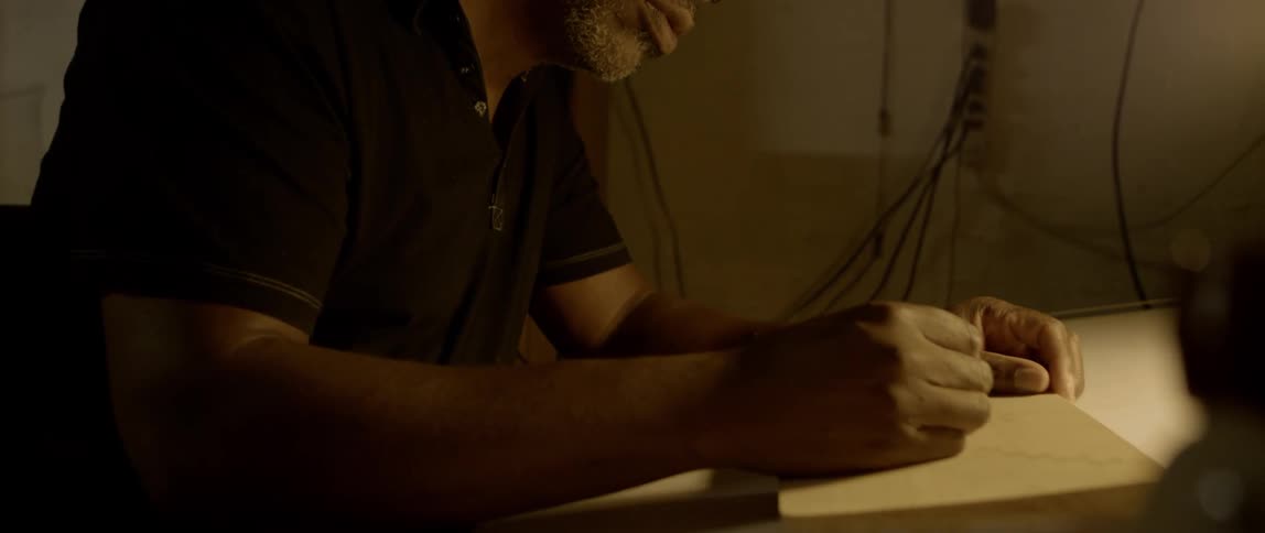 Man sits at his work table illuminated by a small lamp while writing on a square piece of wood using a small pencil