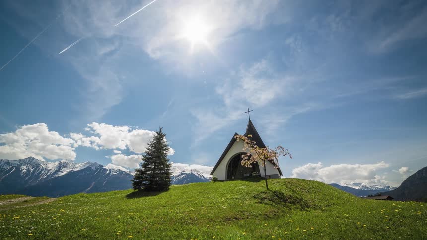 On a bright and sunny day a small church sits atop a grass covered hill and in the distance towering snowcapped mountains can be seen