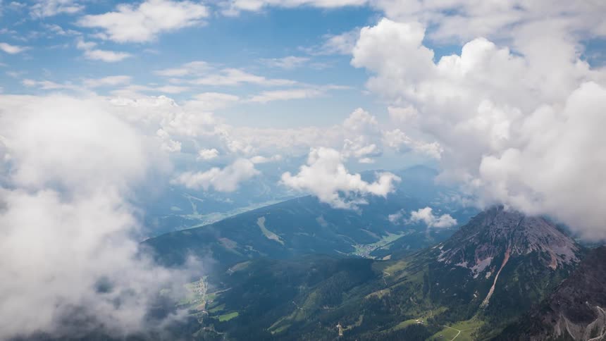 White fluffy clouds are passing above a range of mountains covered in lush green grass and dense trees