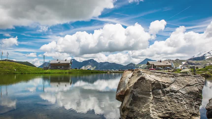 Clouds in a blue sky are reflected on the water surface as people walk around the lake